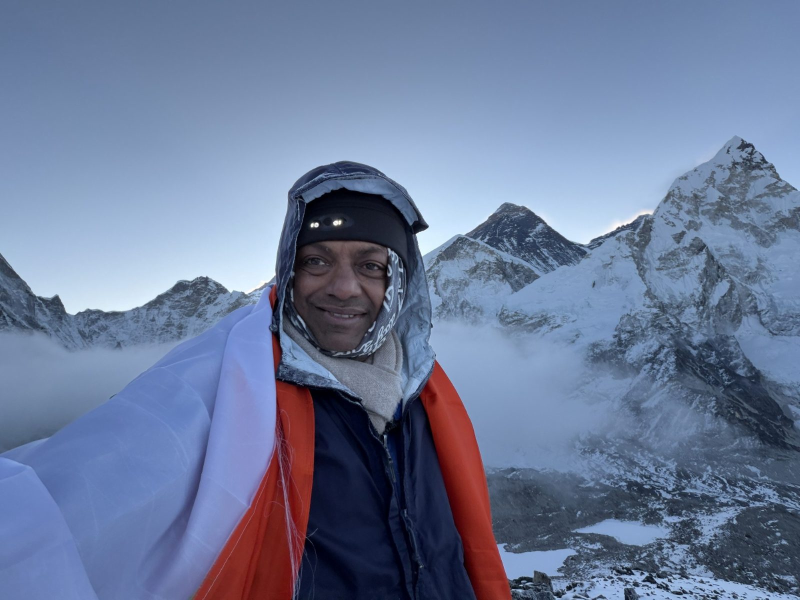 Anand wrapped in a flag on Kala Patthar at dawn, with the dark summit of Mount Everest and the snowy face of Nuptse behind him